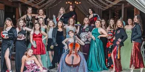 A group of high school seniors posing for a formal portrait in a rustic venue, holding sports equipment and a cello.