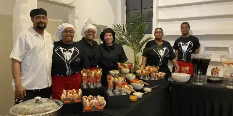Catering staff in uniform standing behind a buffet table with prepared food and chafing dishes