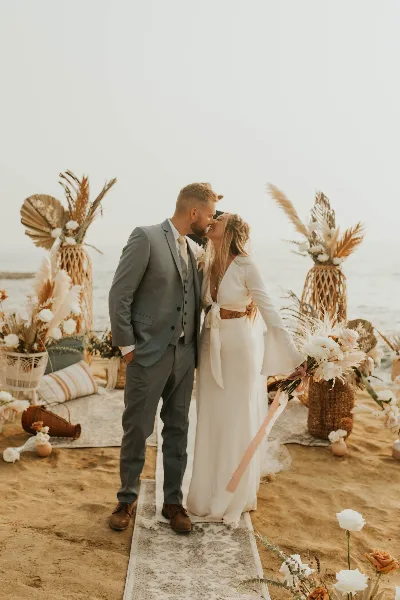 A bride and groom kissing on a beach during a wedding ceremony, surrounded by floral arrangements and event decor.