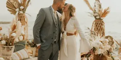 A bride and groom kissing on a beach during a wedding ceremony, surrounded by floral arrangements and event decor.