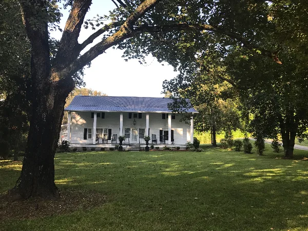 White historic farmhouse with a covered front porch and metal roof set on a grassy lawn framed by large trees.