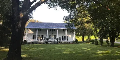 White historic farmhouse with a covered front porch and metal roof set on a grassy lawn framed by large trees.