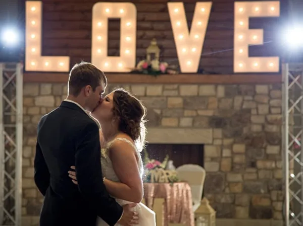 A bride and groom sharing a kiss in front of a large illuminated 'LOVE' sign at a wedding venue.