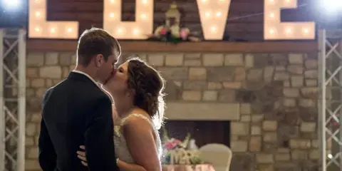 A bride and groom sharing a kiss in front of a large illuminated 'LOVE' sign at a wedding venue.