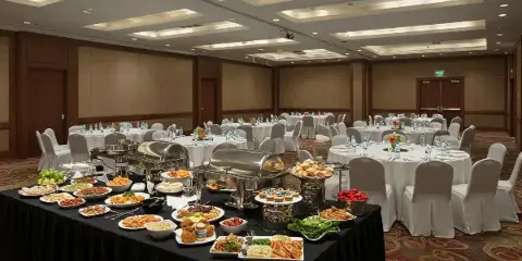 A catering buffet table with chafing dishes and various food items set up in a banquet hall with round tables.