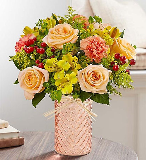 A floral arrangement featuring peach roses, pink carnations, and yellow alstroemeria in a textured copper vase.