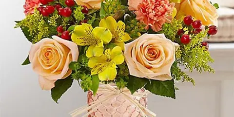 A floral arrangement featuring peach roses, pink carnations, and yellow alstroemeria in a textured copper vase.