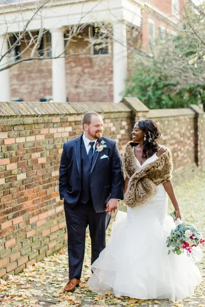 A bride and groom walking hand-in-hand outdoors near a brick wall, with the bride wearing a white gown and fur stole.