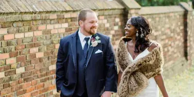A bride and groom walking hand-in-hand outdoors near a brick wall, with the bride wearing a white gown and fur stole.