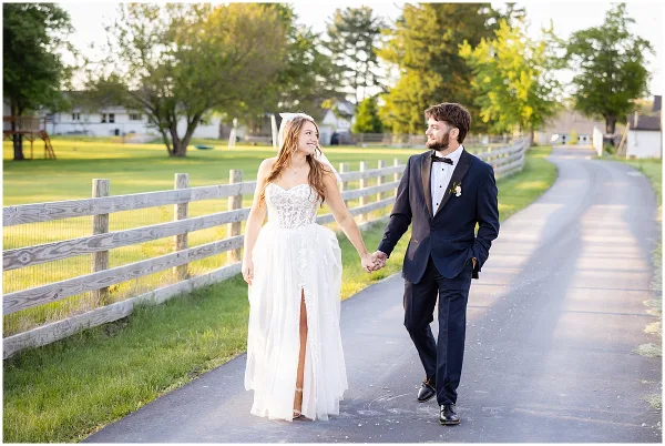 A bride and groom walking hand-in-hand on a path next to a wooden fence in a rural setting.