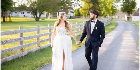 A bride and groom walking hand-in-hand on a path next to a wooden fence in a rural setting.