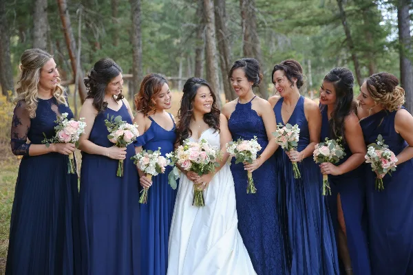 A bride and her bridesmaids in navy blue dresses holding pink and white floral bouquets in a forest setting.