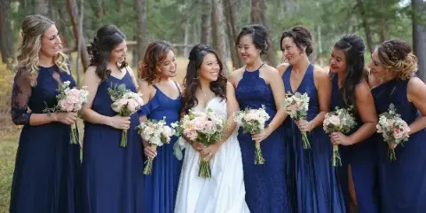 A bride and her bridesmaids in navy blue dresses holding pink and white floral bouquets in a forest setting.