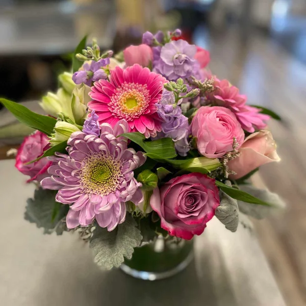 A fresh bouquet of pink and purple flowers, including roses, chrysanthemums, and lilies, arranged in a glass vase.