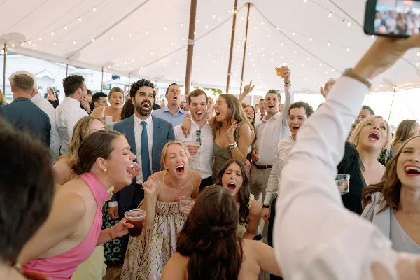 Wedding guests cheering and laughing together under a tent with string lights during a reception.