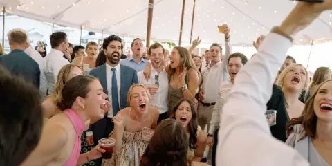 Wedding guests cheering and laughing together under a tent with string lights during a reception.