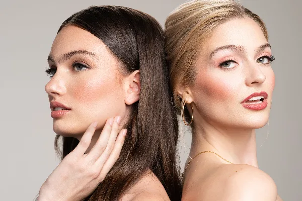 Two women with professional makeup and styled hair posing back-to-back.