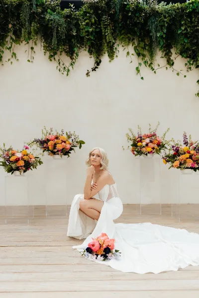 A bride in a white dress kneeling on a wooden floor with a colorful floral bouquet, surrounded by matching floral arrangements on clear pedestals against a white wall with hanging greenery.