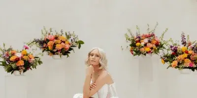A bride in a white dress kneeling on a wooden floor with a colorful floral bouquet, surrounded by matching floral arrangements on clear pedestals against a white wall with hanging greenery.