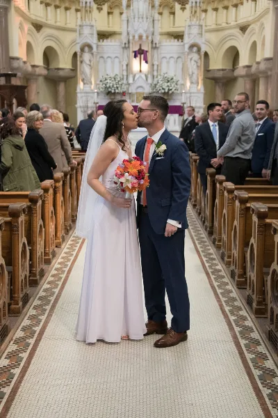 A bride and groom kissing in the aisle of a church during their wedding ceremony, surrounded by guests.