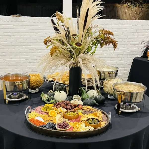 A catering table set with chafing dishes, a large charcuterie board, and a tall floral arrangement.
