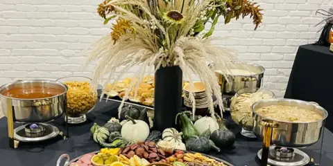 A catering table set with chafing dishes, a large charcuterie board, and a tall floral arrangement.