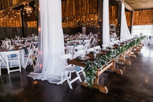 Interior of a wooden-walled wedding reception hall with set tables, white chairs, and string lights.