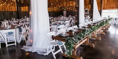Interior of a wooden-walled wedding reception hall with set tables, white chairs, and string lights.
