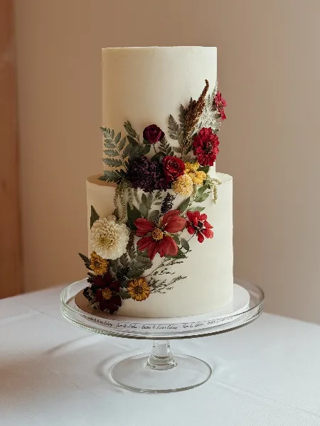 A two-tier white wedding cake decorated with red and yellow flowers on a glass stand with a Butter & Batter Bakery branded board.