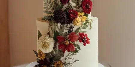 A two-tier white wedding cake decorated with red and yellow flowers on a glass stand with a Butter & Batter Bakery branded board.