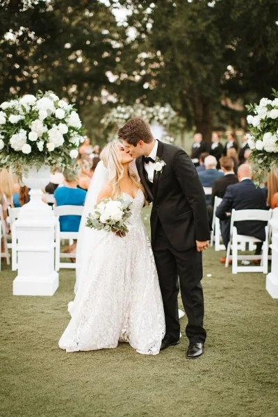 A bride and groom sharing a kiss during an outdoor wedding ceremony, surrounded by white floral arrangements and seated guests.
