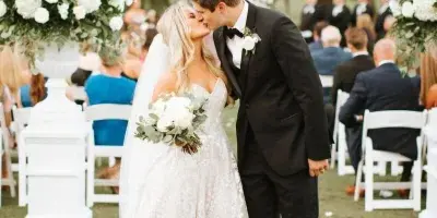 A bride and groom sharing a kiss during an outdoor wedding ceremony, surrounded by white floral arrangements and seated guests.
