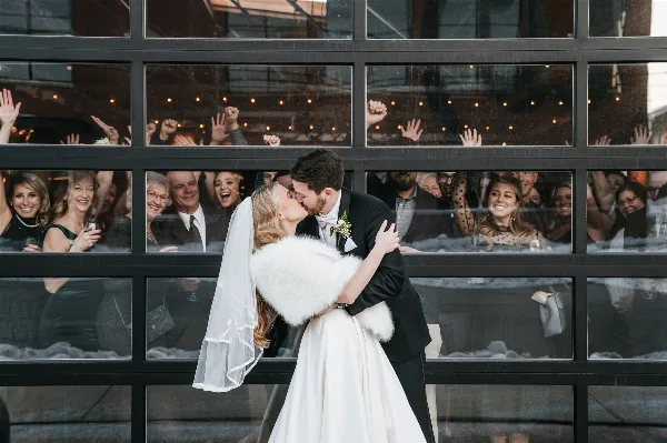A bride and groom kissing in front of a large glass window with wedding guests visible inside the venue.