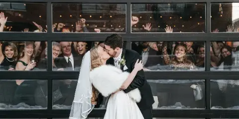 A bride and groom kissing in front of a large glass window with wedding guests visible inside the venue.