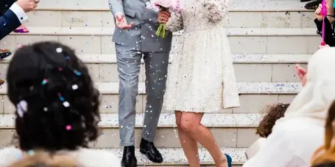 Wedding couple on steps surrounded by guests and falling confetti during a celebration.