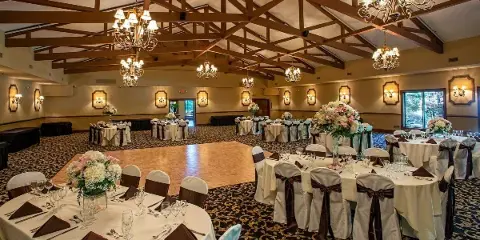 Wedding banquet hall with round tables, floral arrangements, and a central dance floor under a timber-beamed ceiling.