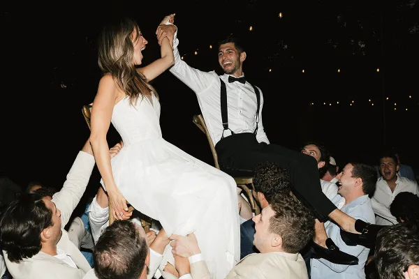 A bride and groom are lifted on chairs by their guests during an outdoor evening wedding reception.
