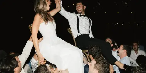 A bride and groom are lifted on chairs by their guests during an outdoor evening wedding reception.