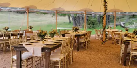 Outdoor wedding reception setup with wooden tables, gold chairs, and floral arrangements under a large tent.