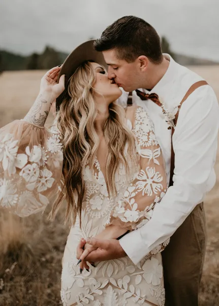A bride and groom sharing a kiss in a field, with the bride wearing a floral lace dress and the groom in a white shirt and suspenders.