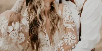 A bride and groom sharing a kiss in a field, with the bride wearing a floral lace dress and the groom in a white shirt and suspenders.