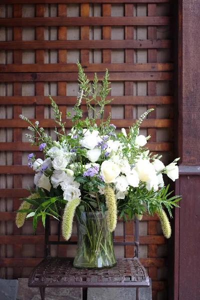 A tall floral arrangement with white roses, purple accents, and greenery in a clear glass vase, set against a wooden lattice background.