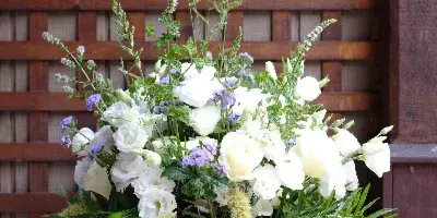 A tall floral arrangement with white roses, purple accents, and greenery in a clear glass vase, set against a wooden lattice background.