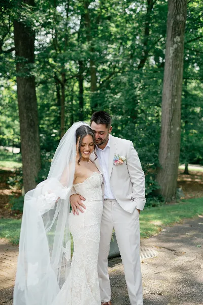 A bride in a lace gown and veil standing with a groom in a cream suit in a wooded area.