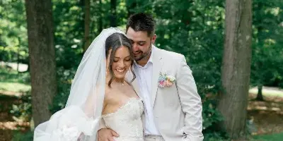 A bride in a lace gown and veil standing with a groom in a cream suit in a wooded area.