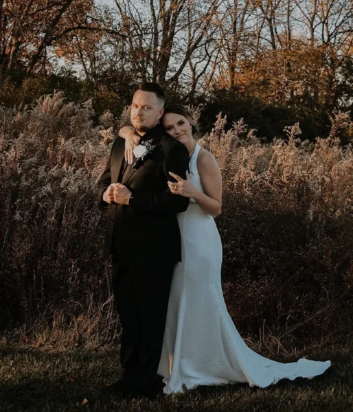 A bride and groom standing together outdoors in a wooded area, wearing wedding attire.