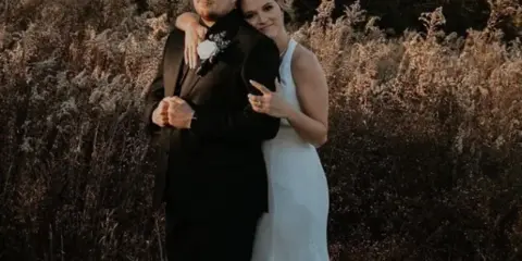 A bride and groom standing together outdoors in a wooded area, wearing wedding attire.