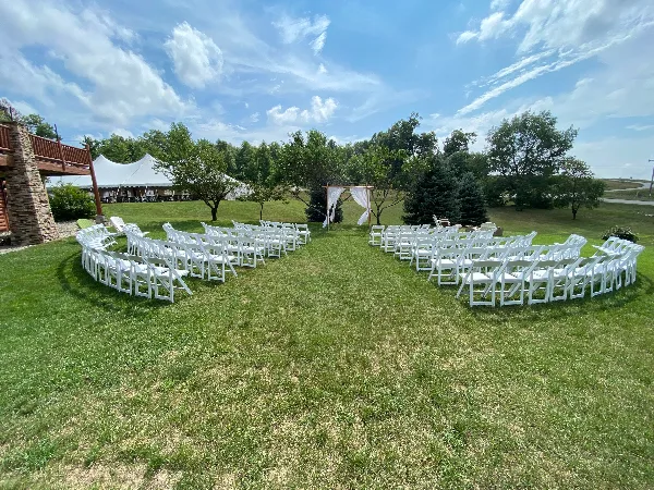 Outdoor wedding ceremony setup with rows of white chairs, a floral arch, and a white tent in the background.