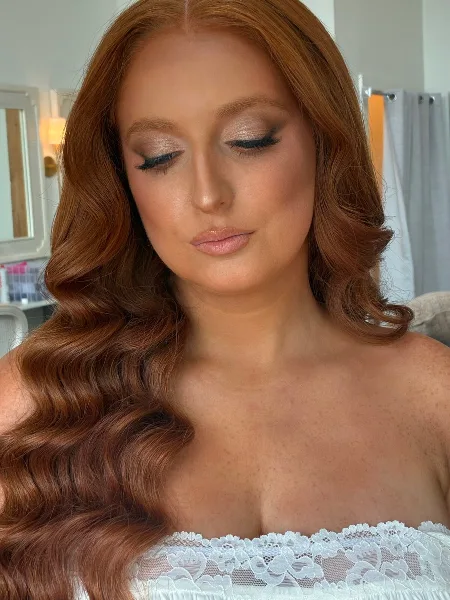 A bride with long, wavy red hair and professional bridal makeup, wearing a white lace wedding dress.