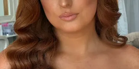 A bride with long, wavy red hair and professional bridal makeup, wearing a white lace wedding dress.